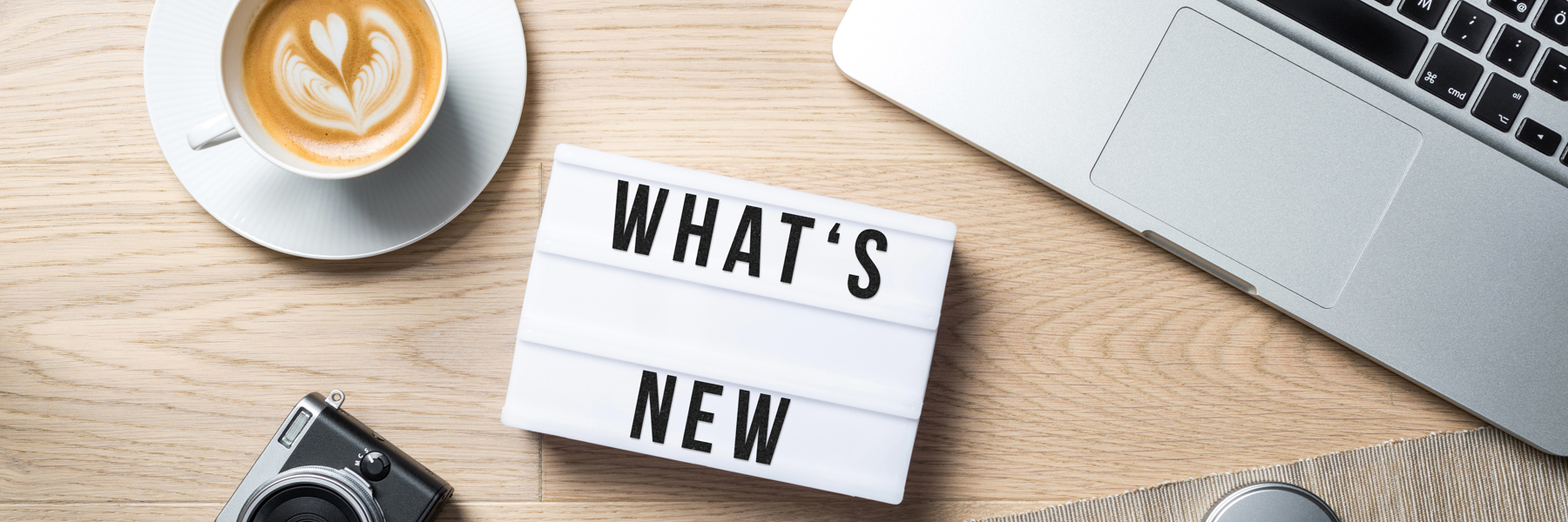 Camera, coffee and ipad open on a desk around a What's new sign