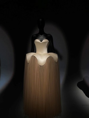 Dark-lit V&A London display of a Schiaparelli strapless gown combining a rigid ivory bodice with soft flowing fringe